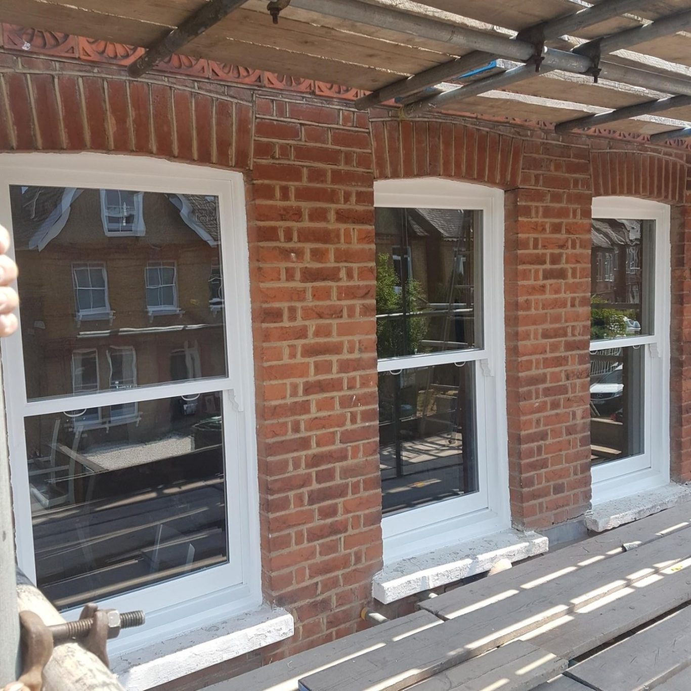 Three white-framed windows on a red brick wall, with scaffolding above.